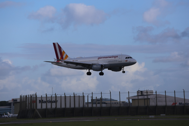 Germanwings Airbus A320-200 beim Start vom Frankfurter Flughafen mit Gras darunter, einem Zaun im Vordergrund, Gebäuden und Bäumen im Hintergrund und einer bewölkten Himmel