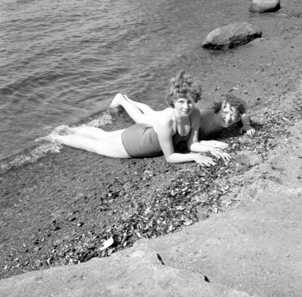 Zwei Frauen in Badesachen liegen auf einem Strand umgeben von Steinen und glitzerndem Wasser, dargestellt in Schwarz-Weiß.