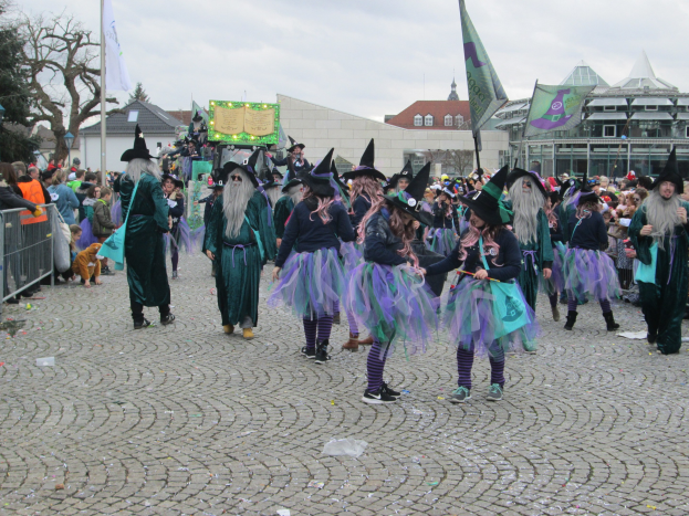 Gruppe von Menschen in Hexenkostümen, die über einen Kopfsteinpflasterstraßen gehen und schwarze und lila Kleider und Hüte tragen, einige halten Fahnen, mit einem Geländer auf der linken Seite und Gebäuden, Bäumen und einem klaren blauen Himmel im Hintergrund.