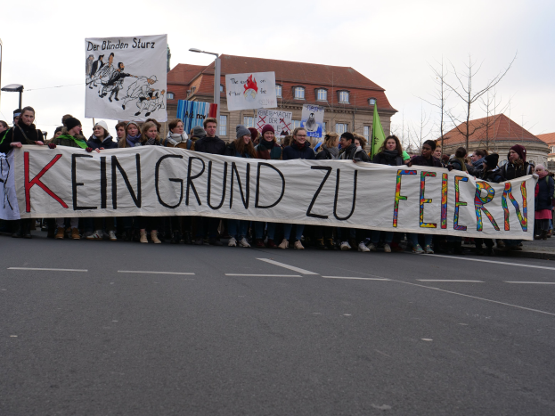 Eine Gruppe von Menschen mit einem Transparent mit der Aufschrift "Kein Grund zu Feiern", das gegen die Sparpolitik der deutschen Regierung protestiert, auf einer Straße mit Laternenpfählen, Straßenlaternen, Bäumen, Gebäuden und einem klaren Himmel.
