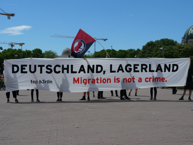 Menschen mit einem Banner, auf dem "Deutschland, Lagerland Migration ist kein Verbrechen" und einer Flagge, vor dem Hintergrund von Straßenlaternen, Bäumen, Kränen, einem Gebäude und einem bewölkten Himmel.