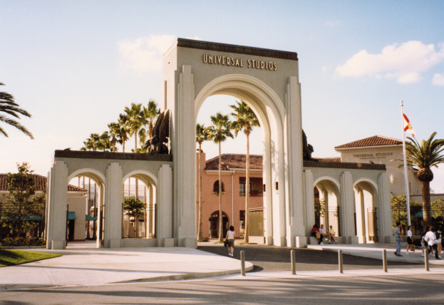 Eingang zum Universal Studios Hollywood mit einem Bogen, einer Statue, Menschen, Bäumen, Gebäuden, einem Fahnenmast und einem bewölkten Himmel.