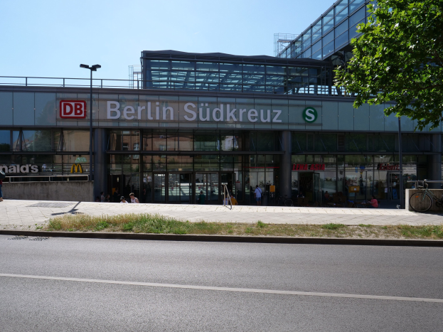 Große gläserne Berliner Südkreuz-Bahnhofgebäude mit Text an seiner Fassade, umgeben von Straßenmasten, Lichtern, Fahrzeugen, Füpßen, Fahrrädern, Bäumen und einem klaren blauen Himmel.