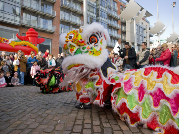 Ein lebendiges chinesisches Neujahrsfest in Amsterdam mit einem Löwen tanzen im Vordergrund und einer Menschenmenge drumherum, vor einem Gebäudehintergrund und einem klaren blauen Himmel, mit einigen Teilnehmern, die das Ereignis fotografieren.