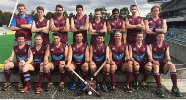 Eine Gruppe von Männern, die auf einem Feld mit Hockeystöcken feiern, umgeben von Bäumen und einem Stadion mit Zuschauern im Hintergrund.