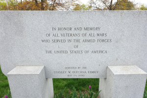 Gedenkstein mit Inschrift für alle US-Veteranen, umgeben von Pflanzen, Blumen, Gras, Bäumen und einer bewölkten Himmel