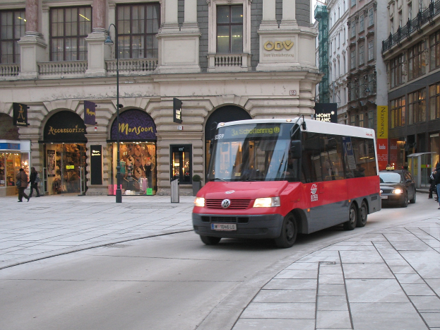 Roter Shuttlebus fährt eine von Hochhäusern gesäumte Stadtstraße entlang, Passanten auf dem Gehweg, Laternenpfähle und Geschäfte mit sichtbarer Beschriftung, mit dem Himmel im Hintergrund.
