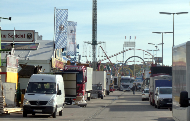 Vollgestaute Straße mit Lastwagen, Autos, Fußgängern, Laternen, Plakaten, einem Turm, einer Attraktion, Bäumen und einem bewölkten Himmel.