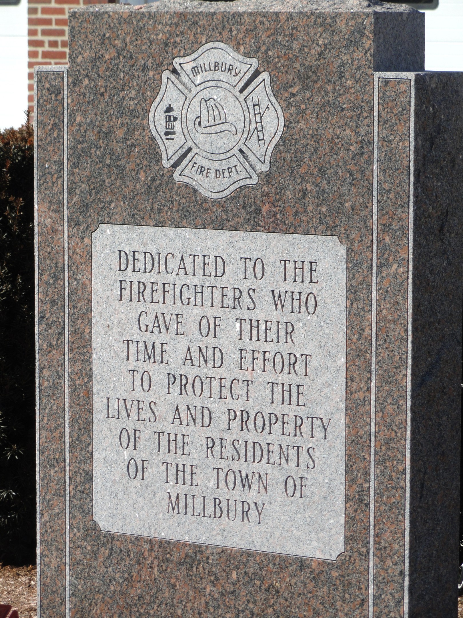 Gedenkstein mit einer Plakette für Feuerwehrleute in Millbury, vor einem Backsteingebäude mit umliegender Vegetation.