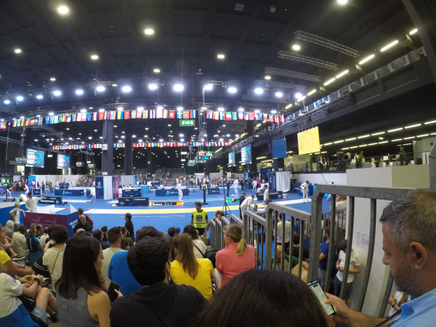 Eine große Menschenmenge sitzt und steht in einer Indoor-Sportarena mit Bildschirmen, Flaggen und Deckenbeleuchtung im Hintergrund.