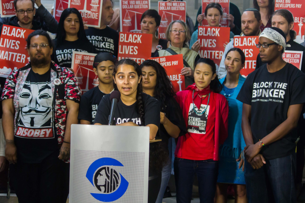 Eine Gruppe von Menschen mit "Black Lives Matter"-Schildern, eine Frau spricht an einem Podium vor einer Menge, vor einer Wand.