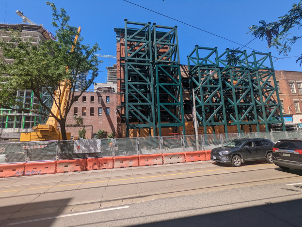 Ein Baustellenszenario mit einer teilweise errichteten Metallbrücke im Vordergrund, Autos, die entlang der Straße geparkt sind, ein Zaun, Bäume, Gebäude mit Fenstern, ein Kran und ein Himmel im Hintergrund.