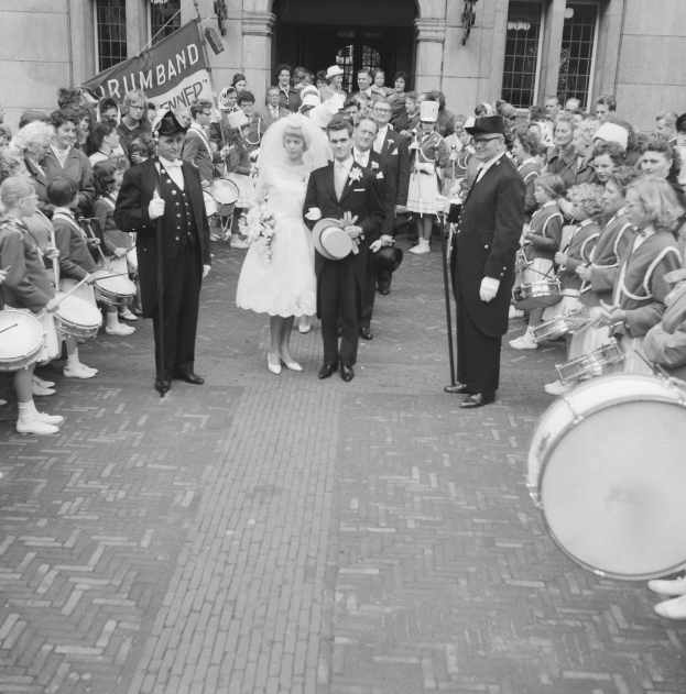 Schwarzes und weißes Foto einer Braut in einem weißen Kleid und Schleier und Bräutigam mit einem Strauß Blumen, der die Straße entlang geht, vorbei an einer Menge, einige halten Musikinstrumente, mit einem Banner und Gebäude im Hintergrund.
