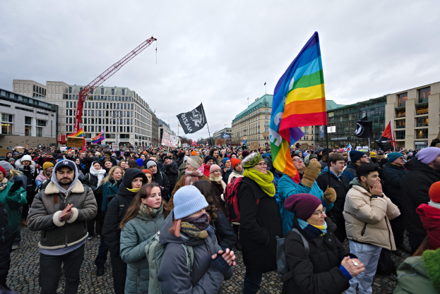 Eine große Gruppe von Menschen bei einer LGBTQ+-Rechtsdemo in Berlin, die Fahnen und Plakate schwenken, mit Gebäuden und einem Kran im Hintergrund.