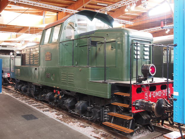 Ein grüner Zugmotor ist auf Schienen in einem Gebäude positioniert, mit einer Plattform auf der linken Seite und einer beleuchteten Decke darüber.