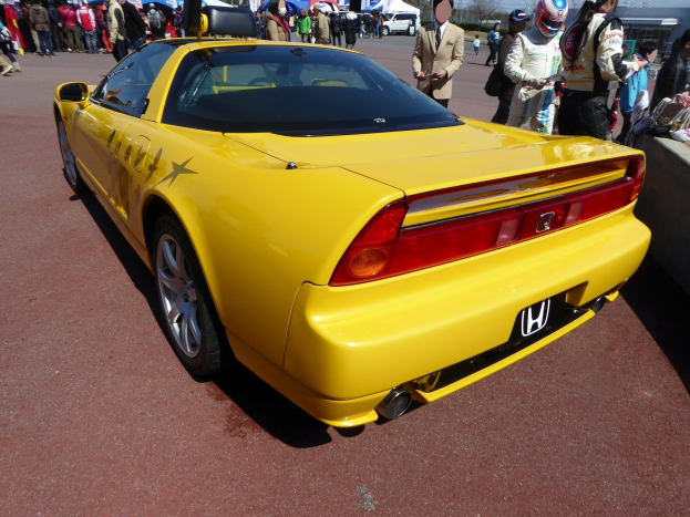 Ein gelber Sportwagen vor einer Menschenmenge geparkt, einige tragen Helme, eine Wand rechts, Zelte, Bäume und Gebäude im Hintergrund bei klarem blauem Himmel.