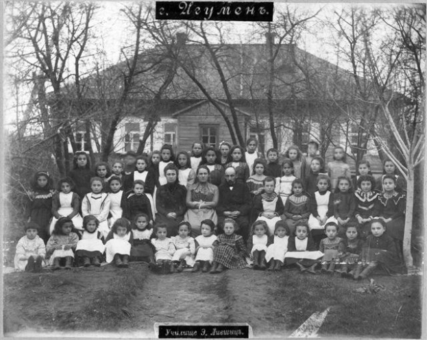 Schwarzes und weißes Foto von Kindern, die vor einem Gebäude mit Fenstern posieren, umgeben von Bäumen und einem klaren Himmel, mit Text unten, der lautet: "Jugoslawien, die erste Schule der Welt."