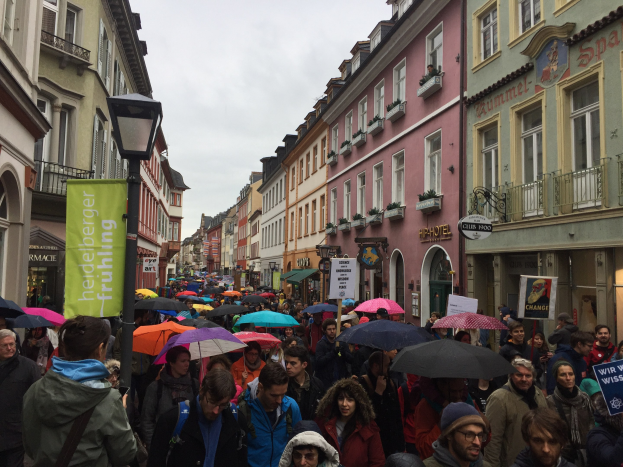 Große Gruppe von Menschen, die mit Schirmen auf der Straße gehen, einige tragen Taschen und halten Schilder, mit Gebäuden und einem Lichtmast im Hintergrund.