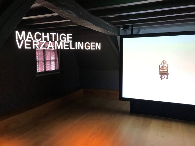 Installationsansicht der Ausstellung "Machtige Verzamelingen" im Museum of Modern Art in Berlin, mit einem großen Bildschirm mit Text an der Wand rechts und einem Fenster links.