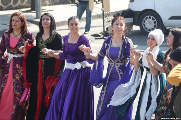Eine Gruppe von Frauen in bunten Kleidern tanzt auf der Straße, im Hintergrund hält jemand eine Kamera, auf der Straße fährt ein Fahrzeug und im Hintergrund ist eine Wand mit Pfählen und anderen Gegenständen zu sehen. Unten steht ein Text, der darauf hinweist, dass sie an einem traditionellen iranischen Tanz teilnehmen.