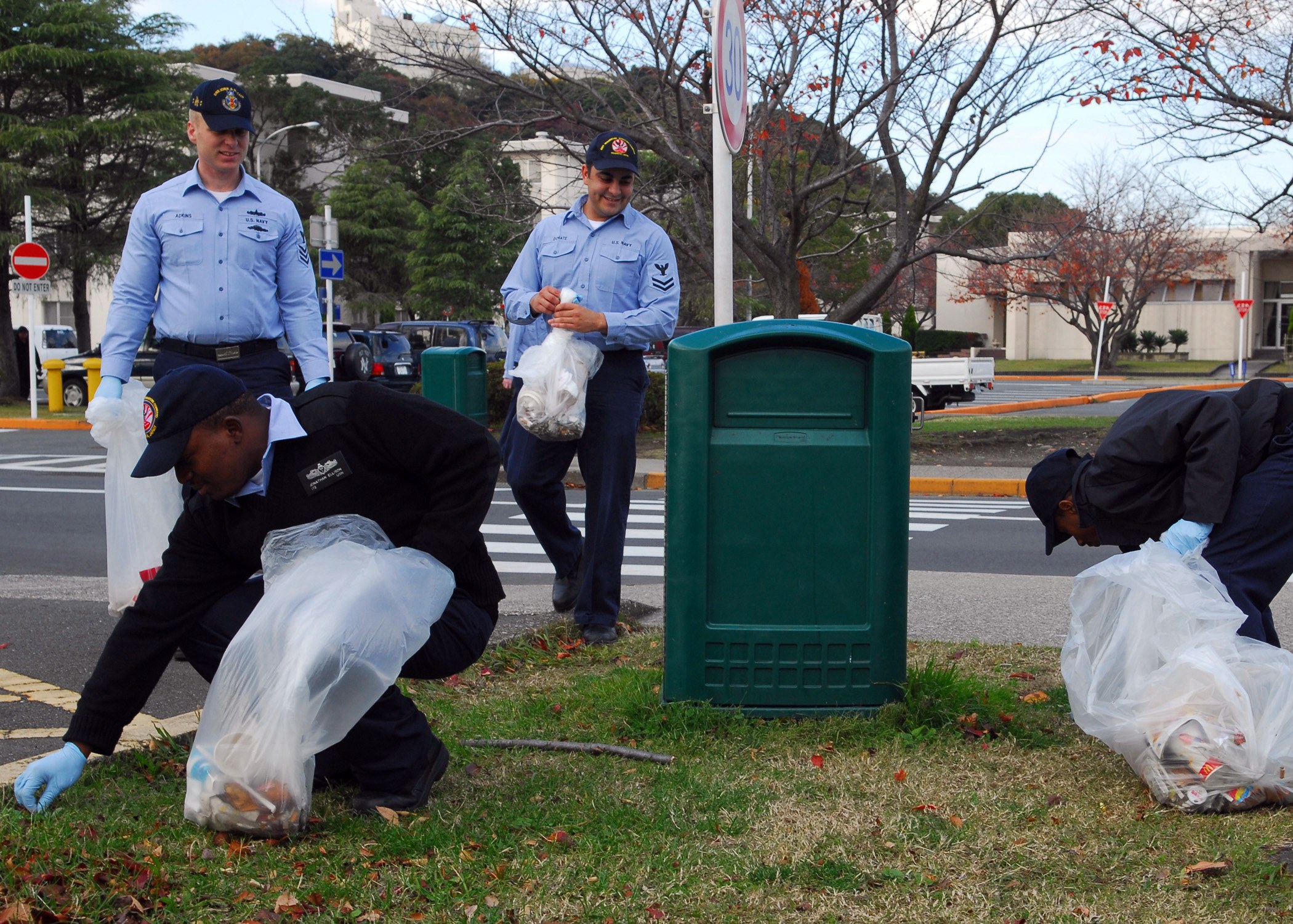 Drei Polizisten in Uniform sammeln Müll am Straßenrand, mit Bäumen, Gebäuden und Fahrzeugen im Hintergrund.