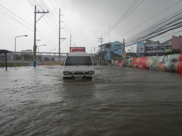 Ein weißer Lieferwagen fährt durch eine von trübem Wasser überschwemmte Stadtstraße, umgeben von Strommasten, Laternen, Gebäuden und einer Brücke im Hintergrund bei klarem Himmel.