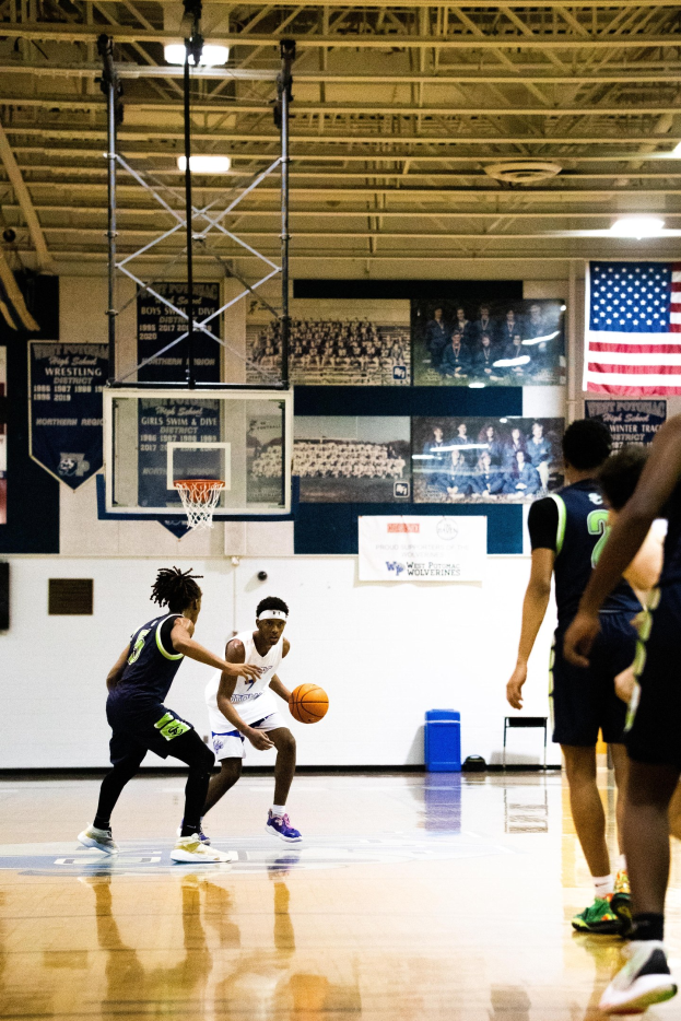 Junge Männer spielen Basketball in einer Halle mit Wand, Deckenstangen und Lampen sowie einer Flagge auf der rechten Seite.