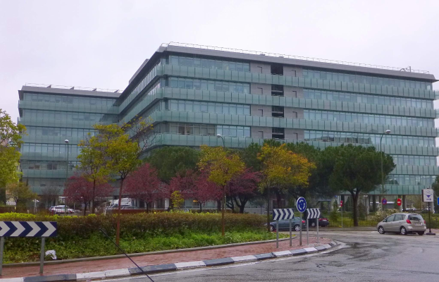 Außenansicht des Hauptsitzes der Europäischen Zentralbank, ein großes Gebäude mit vielen Fenstern, umgeben von Grünflächen und städtischen Elementen wie Straßenlaternen und Fahrzeugen, unter einem bewölkten Himmel.