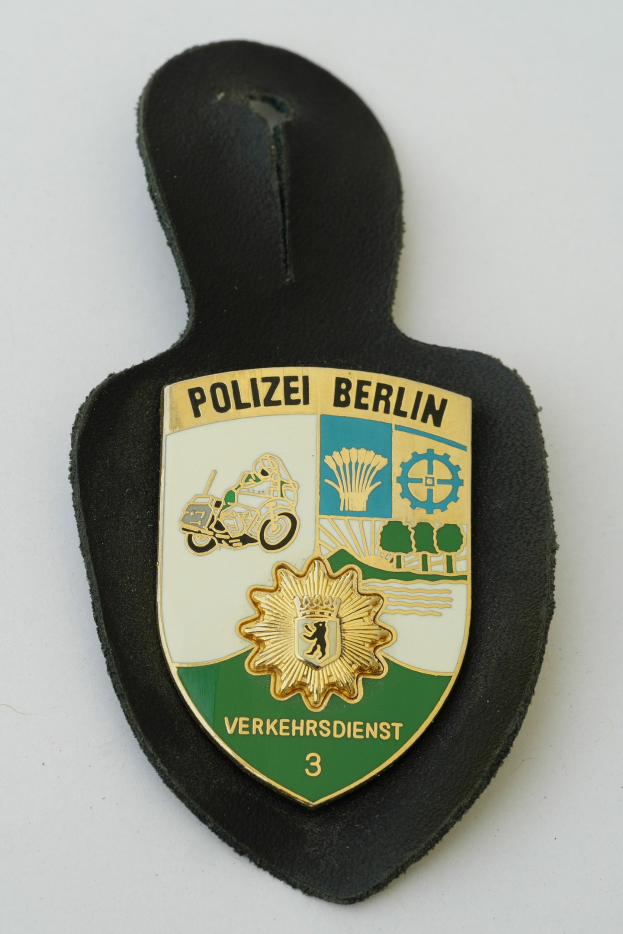 Ein schildförmiges Polizeibadgebnis mit "Polizei Berlin" in schwarzer Schrift, das ein weißes Stern auf einem blauen Hintergrund und einen weißen Rand hat und auf einer Oberfläche ruht.