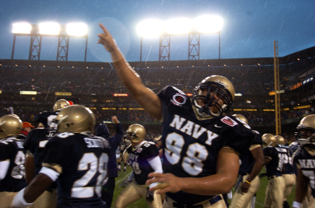 Navy-Football-Spieler feiern Touchdown bei strömendem Regen auf einem grasbewachsenen Feld, mit Helmen und Uniformen, Zuschauern und Stadionbeleuchtung im Hintergrund.