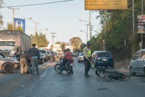 Eine Gruppe von Menschen steht um ein verunglücktes Motorrad auf der Straße mit mehreren Fahrzeugen, darunter ein Lastwagen, und einem Hintergrund aus Bäumen, Pfosten, Lampen und Schildern unter dem Himmel.