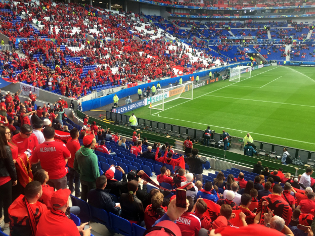 Eine große Menschenmenge in einem Stadion schaut einem Fußballspiel zu, mit Sitzplätzen und Stehplätzen, Fahnen, einem Torpfosten und Deckenleuchten.