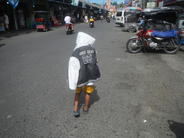 Ein junger Junge geht eine Straße voller Motorräder und anderer Fahrzeuge entlang, flankiert von Gebäuden mit Schildern, unter einem klaren blauen Himmel mit Bäumen und Masten im Hintergrund.
