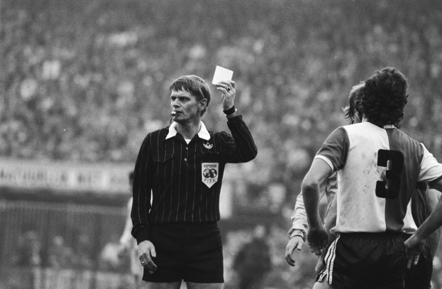 Schwarzes und weißes Bild eines Schiedsrichters mit roter Karte, mit einer Menge von Menschen in Fußballtrikots hinter einem Zaun.