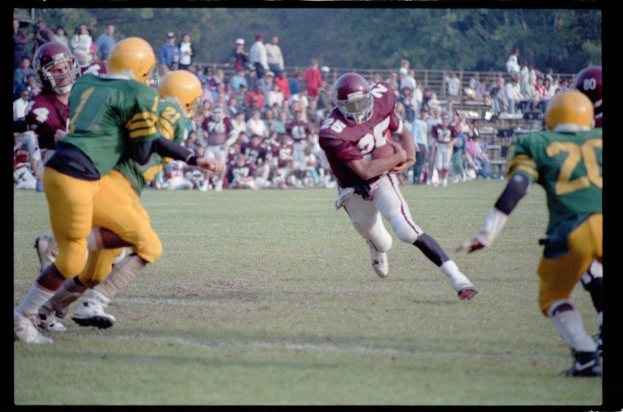Fußballspieler in Bewegung mit dem Ball, umgeben von Gegenspielern und Mannschaftskameraden in Sportbekleidung, mit Zuschauern und Bäumen im Hintergrund und Text unten.