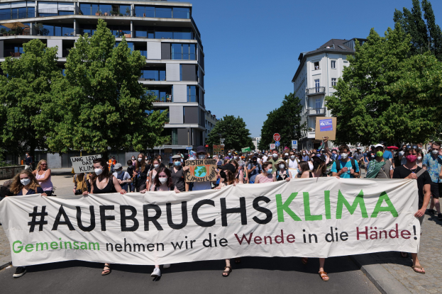 Gruppe von Menschen mit Masken, die eine Fahne mit der Aufschrift 'Aufbruchsklima' vor einem Gebäude mit Bäumen und klarem Himmel halten.