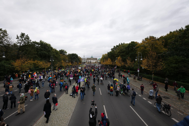 Eine große Gruppe von Menschen marschiert auf einer von Bäumen gesäumten Straße in Berlin, mit Kameras in der Hand, mit einem Gebäude und einem klaren Himmel im Hintergrund.
