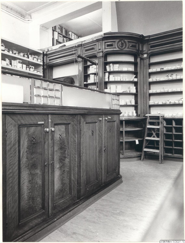 Ein Schwarz-Weiß-Foto eines Apothekeninneren mit einer Theke, Schränken, einer Leiter und Regalen, die mit verschiedenen Gegenständen gefüllt sind, mit Text oben auf dem Bild.