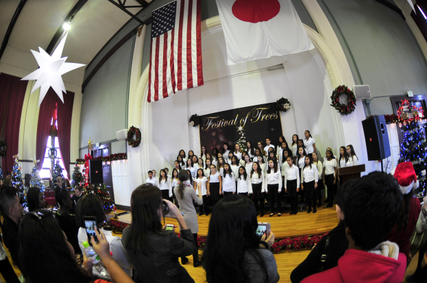 Eine Gruppe von Menschen auf einer Bühne vor einem Publikum mit Weihnachtsdekoration, einem Rednerpult, Lautsprechern, einer Texttafel, Vorhängen, Fahnen und Deckenleuchten.