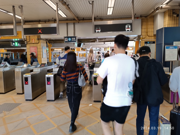 Eine Gruppe von Menschen geht durch einen Bahnhof, einige tragen Taschen, mit Metalldetektoren, Hinweisschildern, Metallstangen und Deckenleuchten sowie einem Wasserzeichen im rechten unteren Eck.