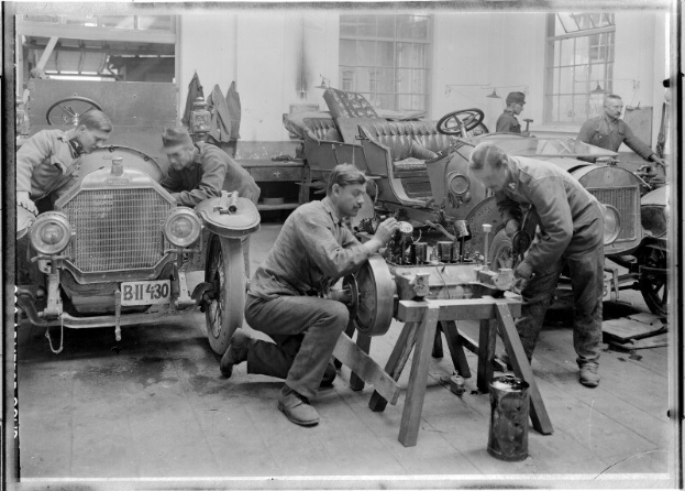 Ein Schwarz-Weiß-Bild von mehreren uniformierten Männern, die ein Auto in einer Garage reparieren, mit Werkzeugen und Equipment auf einem zentralen Tisch.