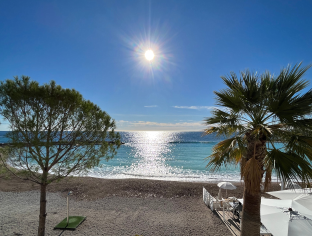 Ein Strand mit Palmen, Sonnenschirmen, üppiger Vegetation, einem strahlend blauen Himmel und einer leuchtenden Sonne im Hintergrund, geeignet für eine Ferienwohnung an der französischen Riviera.