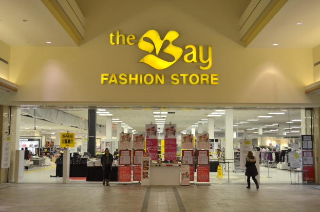 Außenansicht eines Bay Fashion Store in einem Einkaufszentrum mit Menschen, die gehen, Texttafeln an den Wänden, Säulen, Deckenleuchten und Hintergrundauslagen.