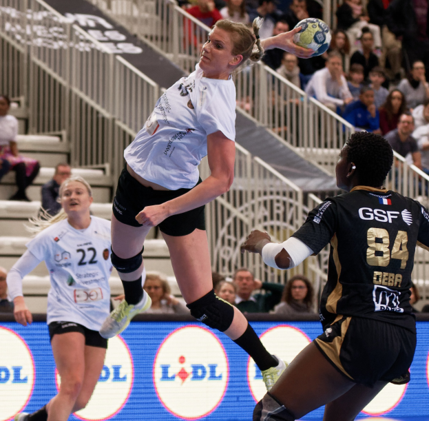 Eine Gruppe von Frauen, die auf einem Platz Handball spielen, mit einem Ball in der Luft, Zuschauern auf Treppen und einer "Handball-Weltmeisterschaft 2019 - Tag 2"-Plakette im Hintergrund.