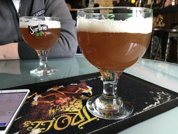 Zwei Gläser goldenes Bier auf einem Tisch neben einem Handy, mit Menschen im Hintergrund und gerahmten Kunstwerken an der Wand.