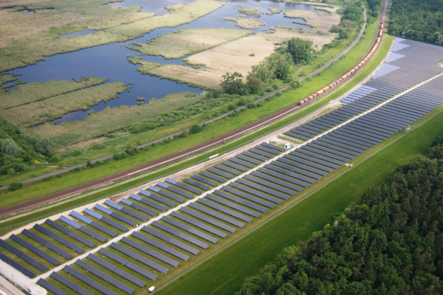 Eine Luftaufnahme eines Solarparks mit zahlreichen Solarmodulen in einem Feld, umgeben von Bäumen, Gras und Wasser, mit einem Zug, der auf einem Bahngeleis in der Nähe fährt.