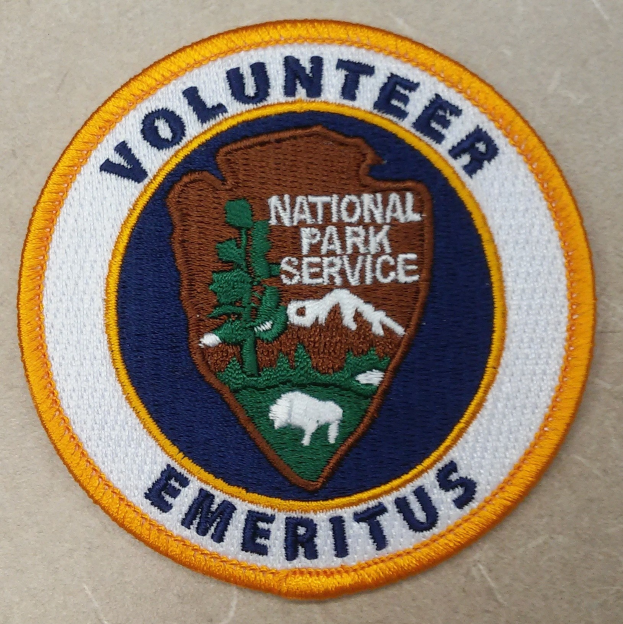Ein kreisförmiges Abzeichen mit einem blauen Rand, weißem Hintergrund und fettem schwarzem Text, der 'Volunteer National Park Service Emeritus' in der Mitte eines gelben Kreises liest.