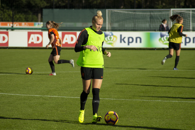 Gruppe von Frauen in Fußballtrikots beim Spiel auf einem Feld mit Bäumen im Hintergrund, Banner hinter ihnen und ein Netz im Vordergrund.