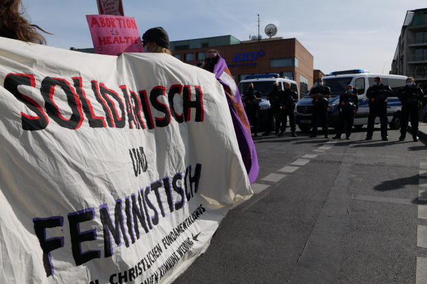 Eine Gruppe von Menschen marschiert auf einer Straße, hält ein Banner mit der Aufschrift "Solidarität und Feminismus" und hat parkende Fahrzeuge und Gebäude im Hintergrund bei klarem blauem Himmel.