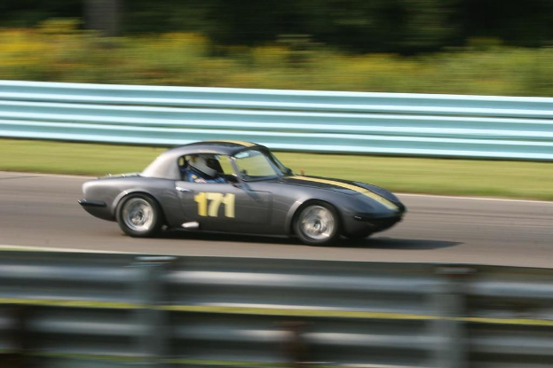 Ein Auto mit einem Helmträger fährt auf einer Straße mit Absperrungen, im Hintergrund sind Bäume zu sehen.
